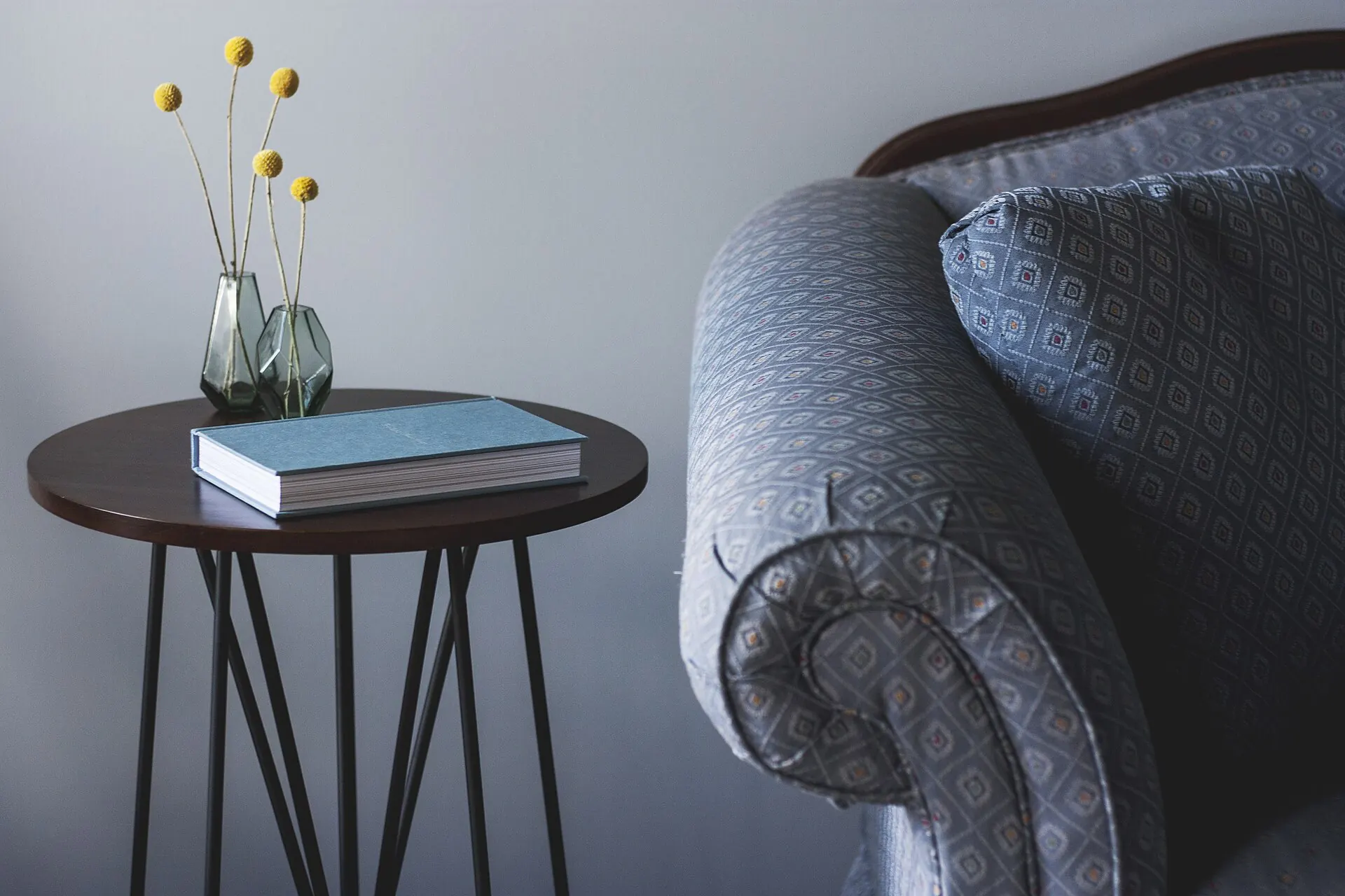 The rolled arm of a grey patterned upholstered sofa beside a small side table